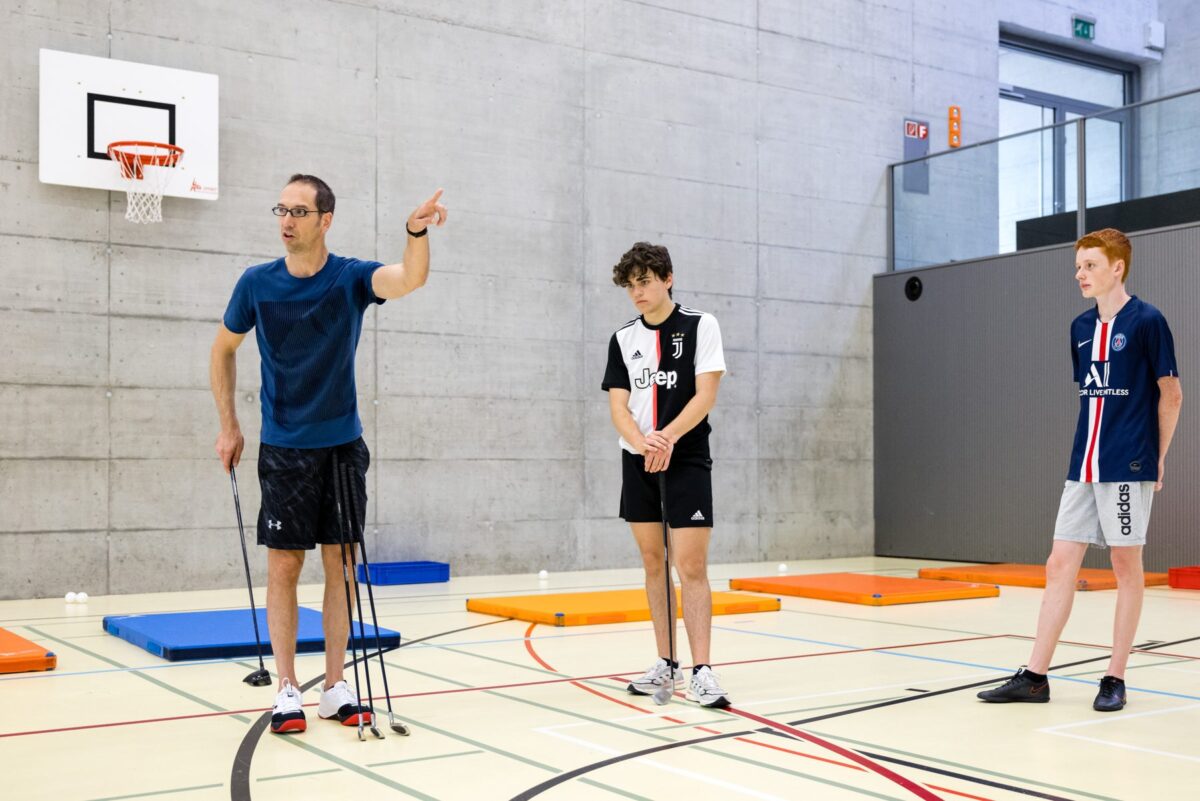 Eine Lehrperson erklärt eine Übung mit Schlägern in der Turnhalle.